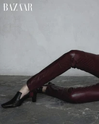 Close-up of burgundy leather trousers and black heeled loafers against a mottled grey wall and textured floor.