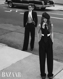 Two models in tailored black and white suits standing on an asphalt street with a vintage car in the background.