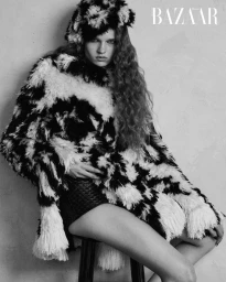 Model in black and white patterned fluffy coat and matching hat seated on a stool against a textured wall.