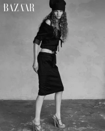 Model in black crop top and skirt wearing a dark hat against a mottled wall