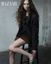 Model with long curly brown hair seated on a stool wearing a black blazer and brown skirt against a rustic concrete wall.