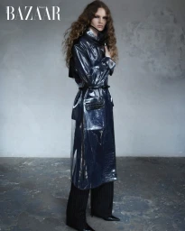 Model in glossy dark vinyl trench coat and dark trousers standing against a textured concrete wall.