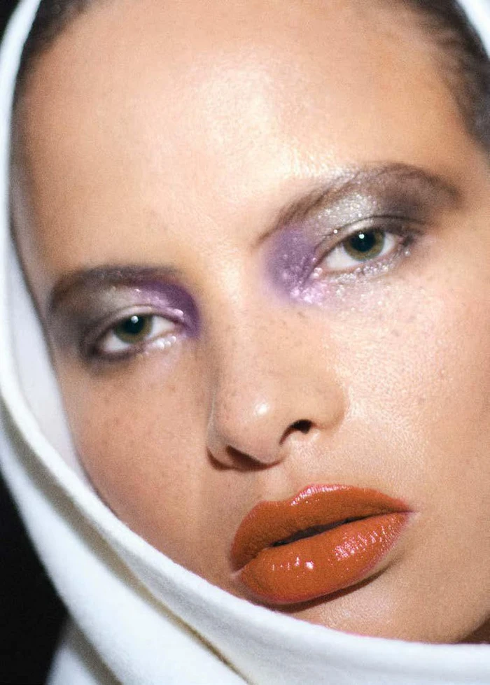 Close-up portrait of a model with glossy terracotta lips and purple eyeshadow framed in a white head covering for Puss Puss Magazine