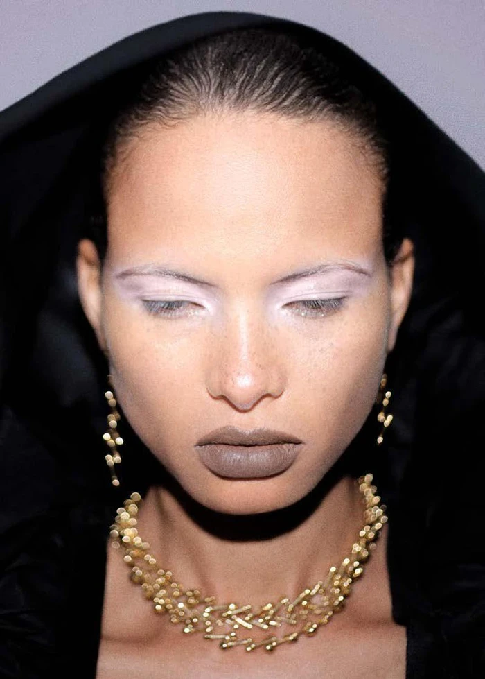 Close-up portrait of model Litza Veloz wearing gold statement jewelry and a black hooded garment for Puss Puss Magazine.