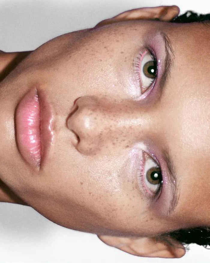 Extreme close-up portrait of a woman with freckles, pink eyeshadow, and glossy lips against a neutral background.