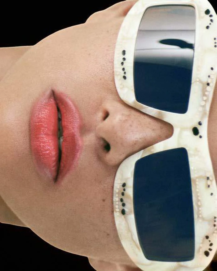 Close-up portrait of a model wearing chunky white sunglasses and pink lipstick against a dark background.