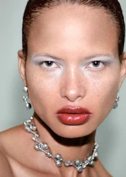 Close-up portrait of a model with glossy red lips, pale eyeshadow, silver necklace, and earrings against a plain background