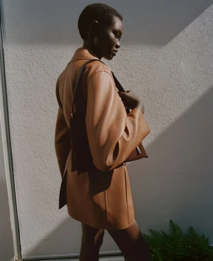 Model Ateta Jok wearing a brown coat and carrying a shoulder bag against a textured exterior wall in Silver Rain project for Puss Puss Magazine