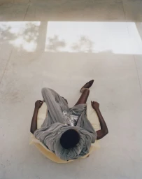 Overhead shot of a model in grey draped clothing seated in a chair on a concrete floor with light patterns.