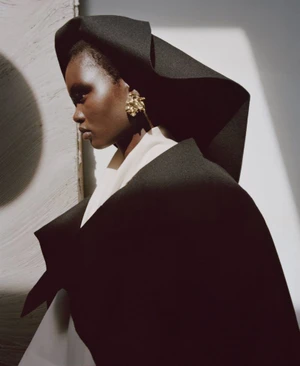 Model Ateta Jok wearing a structured black sculptural coat and headpiece with gold earrings for Puss Puss Magazine