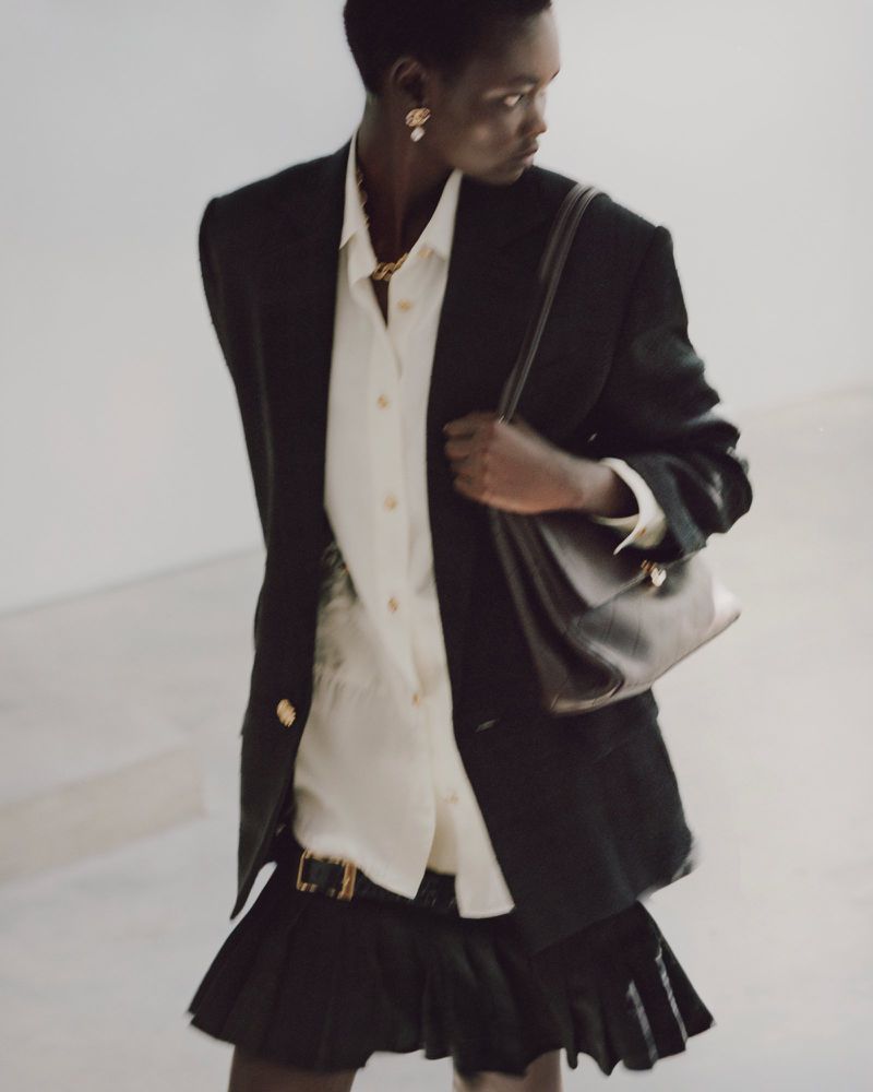 Model wearing a black blazer, white shirt, and dark skirt holding a leather shoulder bag against a white background