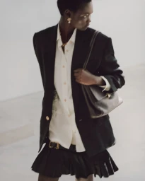 Model wearing a black blazer, white shirt, and dark skirt holding a leather shoulder bag against a white background