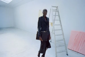 Model Ateta Jok in a dark leather coat holding a dark handbag in a gallery space with a ladder and pink panel