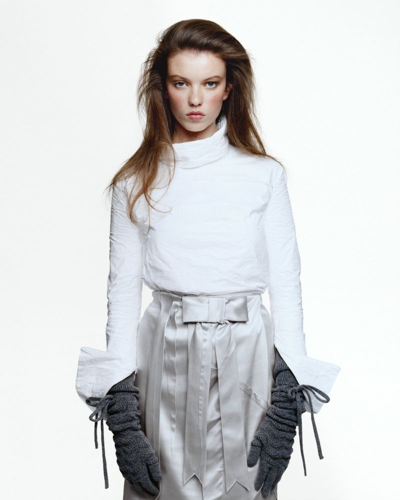 Model wearing a white turtleneck and grey bow skirt with long knitted gloves against a white seamless background