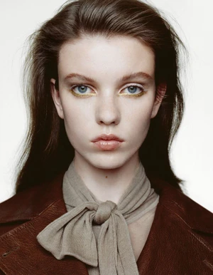 Close-up portrait of a woman with long dark hair, wearing a tied neck scarf and dark jacket against a plain white background.