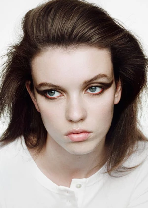 Close-up portrait of a model with dark brown hair and dramatic eye makeup wearing a white top against a white background.