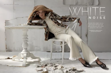 Model seated on a white chair leaning against a white table wearing a patterned cardigan and light trousers