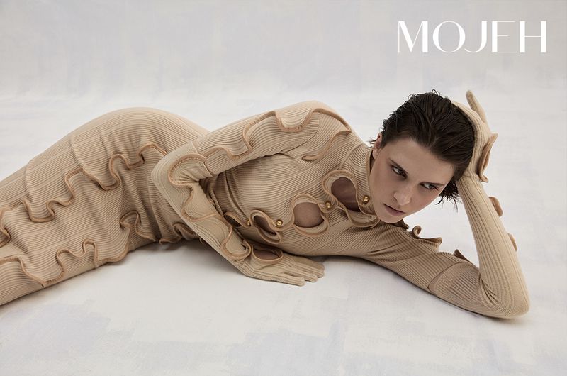 Model lying on a light background wearing a beige textured ribbed dress with cutout details and integrated gloves for Mojeh Magazine.