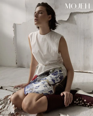 Model seated on a textured white floor wearing a white sleeveless top and patterned skirt for Mojeh Magazine