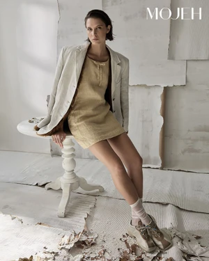 Model seated on a white pedestal table wearing a tan dress, oversized beige blazer, sheer socks, and sneakers.