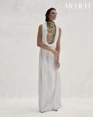 Model in a long white dress and patterned high-neck top standing against a neutral studio background for Mojeh Magazine editorial