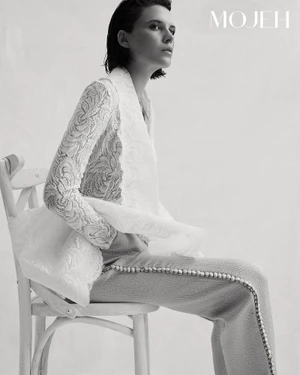 Monochrome fashion editorial image of a model seated on a chair wearing a white patterned lace jacket and trousers