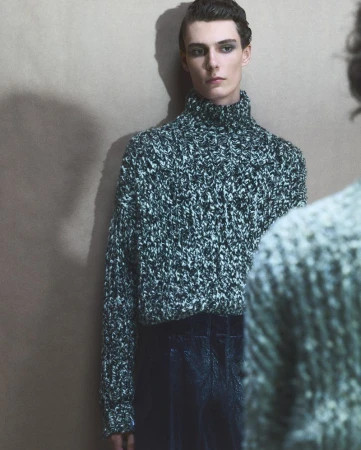 Model in a green turtleneck sweater standing against a plain neutral wall with a shadowed reflection in the foreground