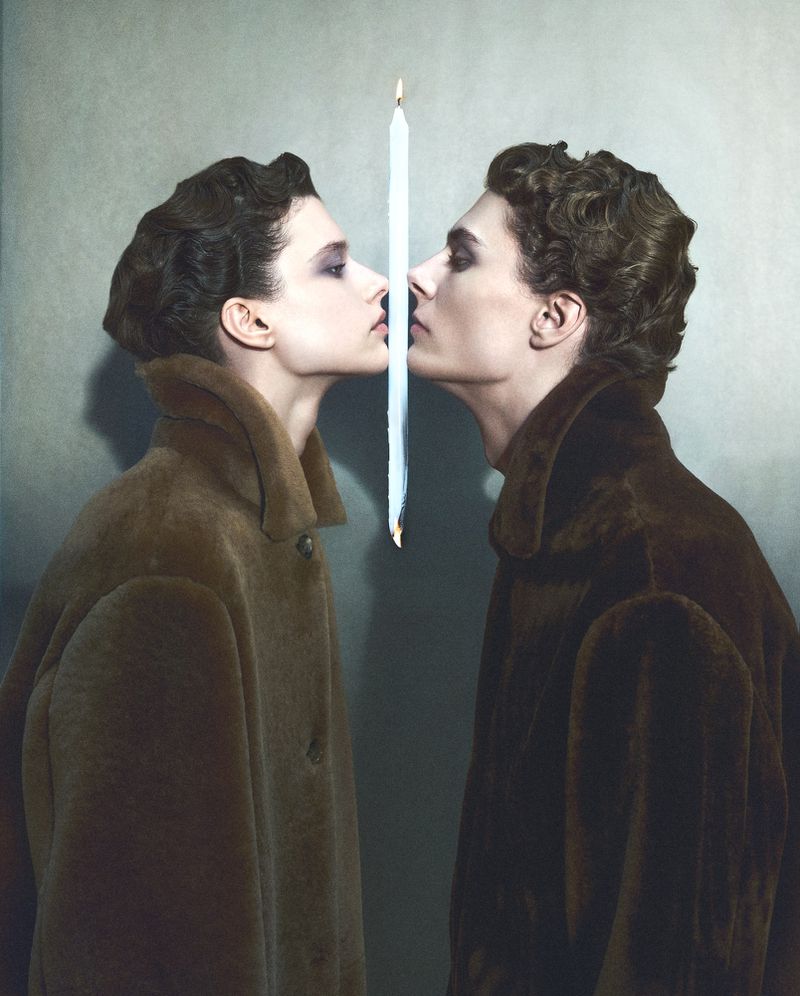 Two models in brown coats facing each other separated by a burning vertical candle