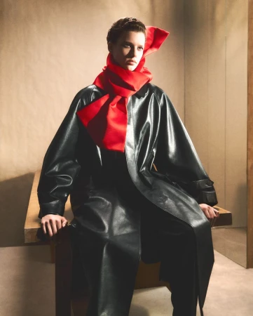 Model in dark leather coat wearing a large red scarf seated on a wooden table against a neutral wall