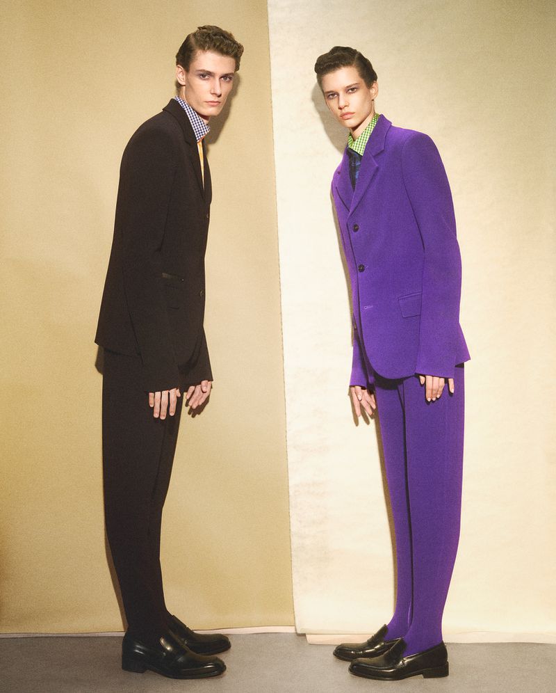 Two models wearing tailored suits standing against a two-tone studio backdrop in an editorial portrait.