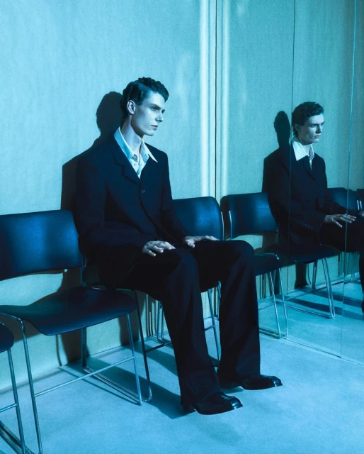 A man in a black suit seated on a chair with a mirror reflection in a cool blue-toned studio setting.
