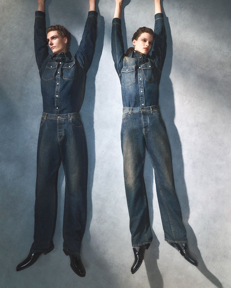 Two models standing against a grey textured wall wearing matching denim shirts and jeans with arms raised.