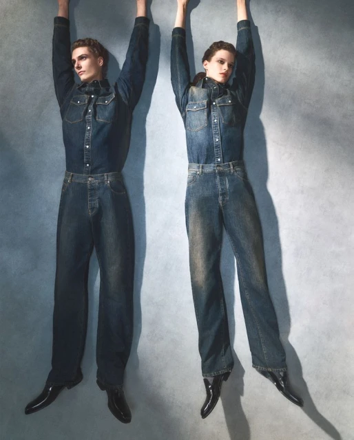 Two models standing against a grey textured wall wearing matching denim shirts and jeans with arms raised.