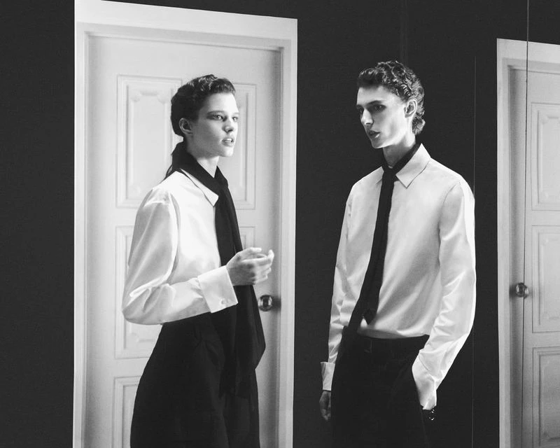 Black and white editorial portrait of two models wearing white shirts and dark ties against a doorway.