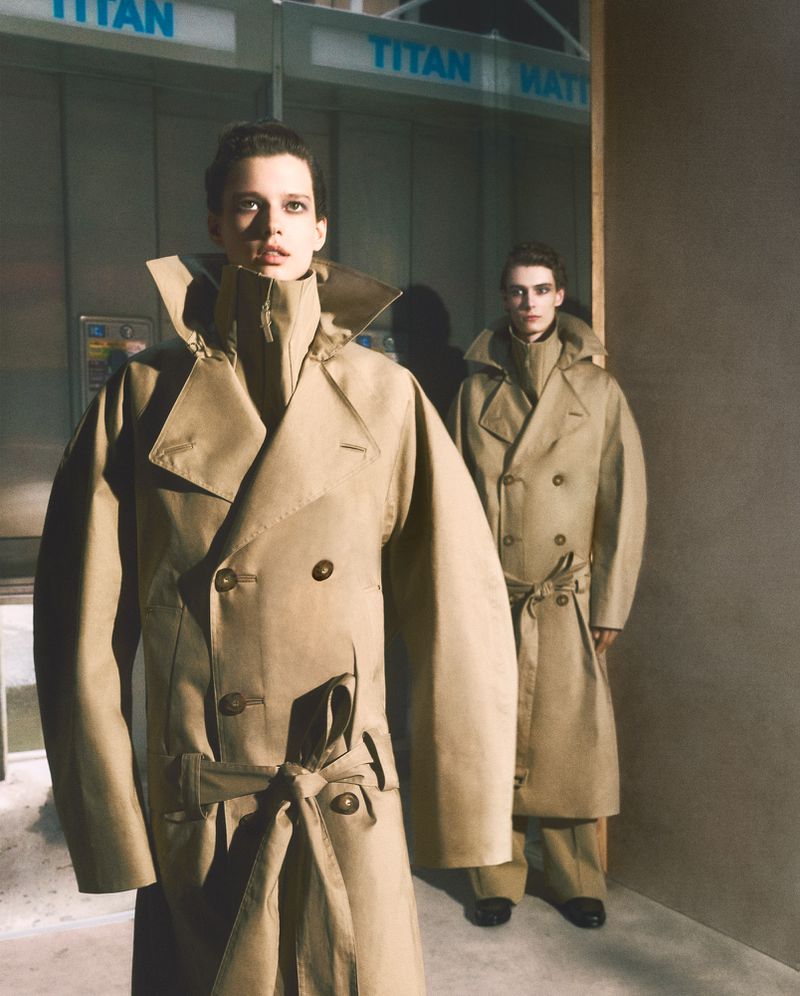 Two models wearing beige double-breasted trench coats standing in an interior space with a telephone booth backdrop