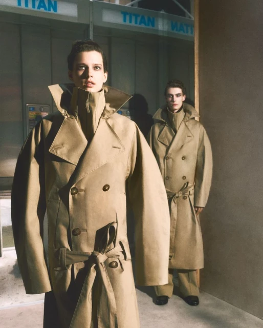 Two models wearing beige double-breasted trench coats standing in an interior space with a telephone booth backdrop