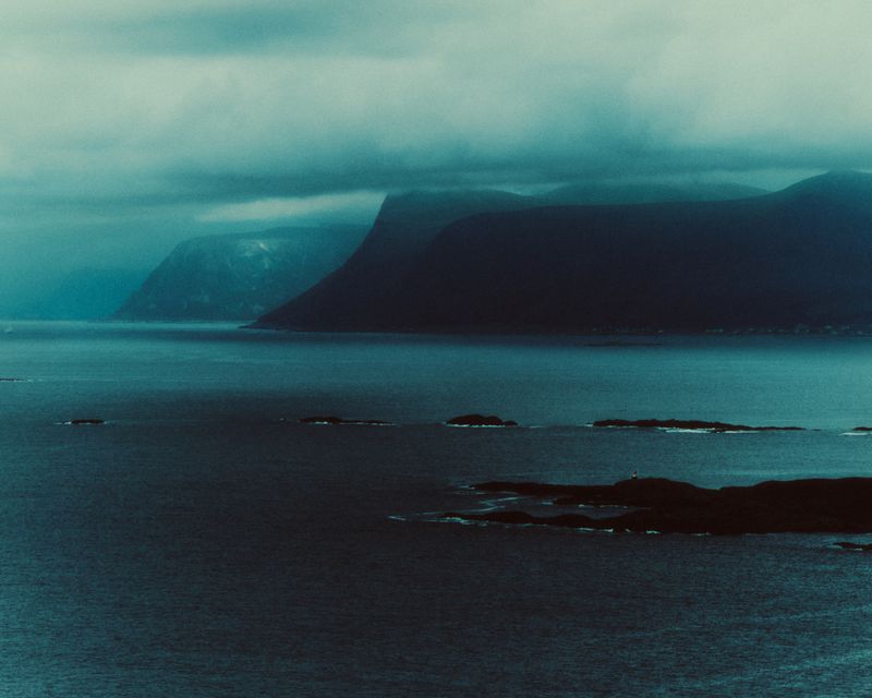 Moody aerial landscape of dark mountainous coastlines bordering deep blue ocean waters under a heavy blanket of low clouds.