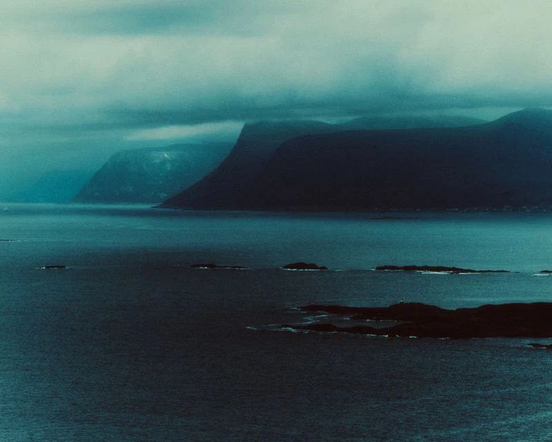 Moody aerial landscape of dark mountainous coastlines bordering deep blue ocean waters under a heavy blanket of low clouds.