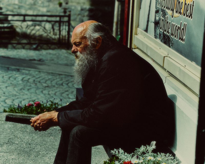 Elderly man with a long white beard seated outdoors on a bench next to a paved stone street.