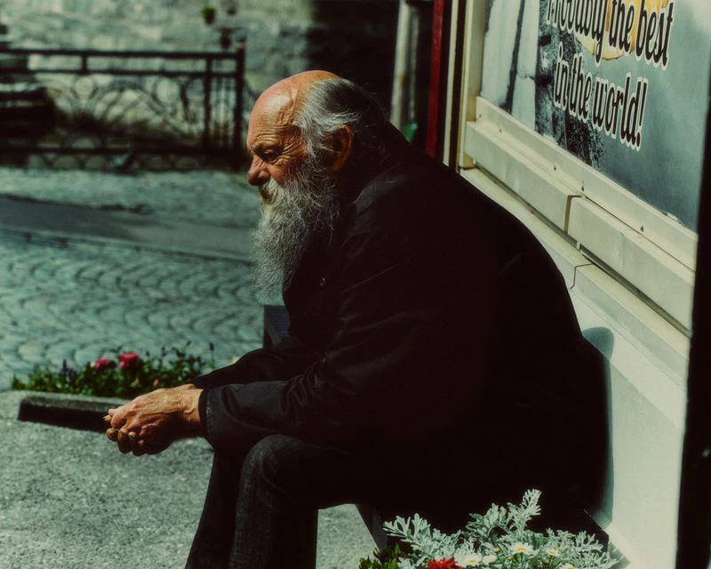 Elderly man with a long white beard seated outdoors on a bench next to a paved stone street.
