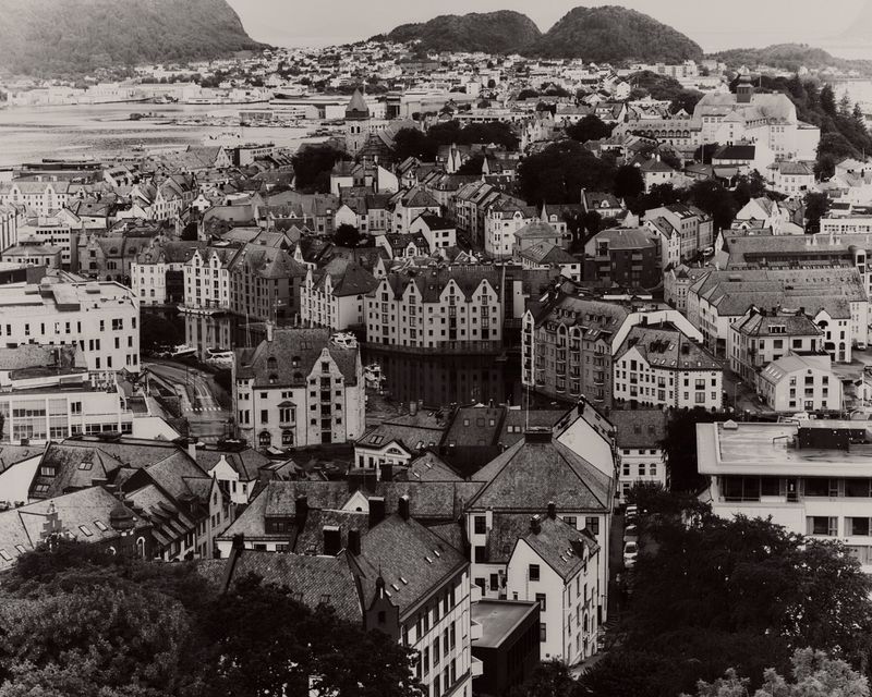 High-angle black and white aerial photograph of a coastal town with densely clustered buildings and rolling hills in the background.