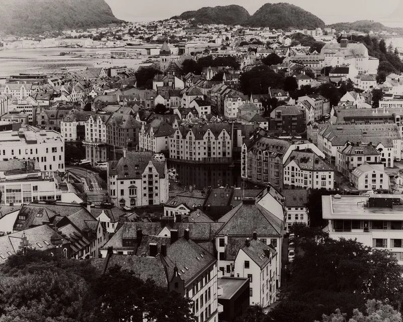 High-angle black and white aerial photograph of a coastal town with densely clustered buildings and rolling hills in the background.