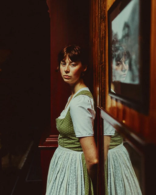 Woman in green dirndl leaning against a wood-paneled wall with a framed picture in a moody interior by Max Miechowski