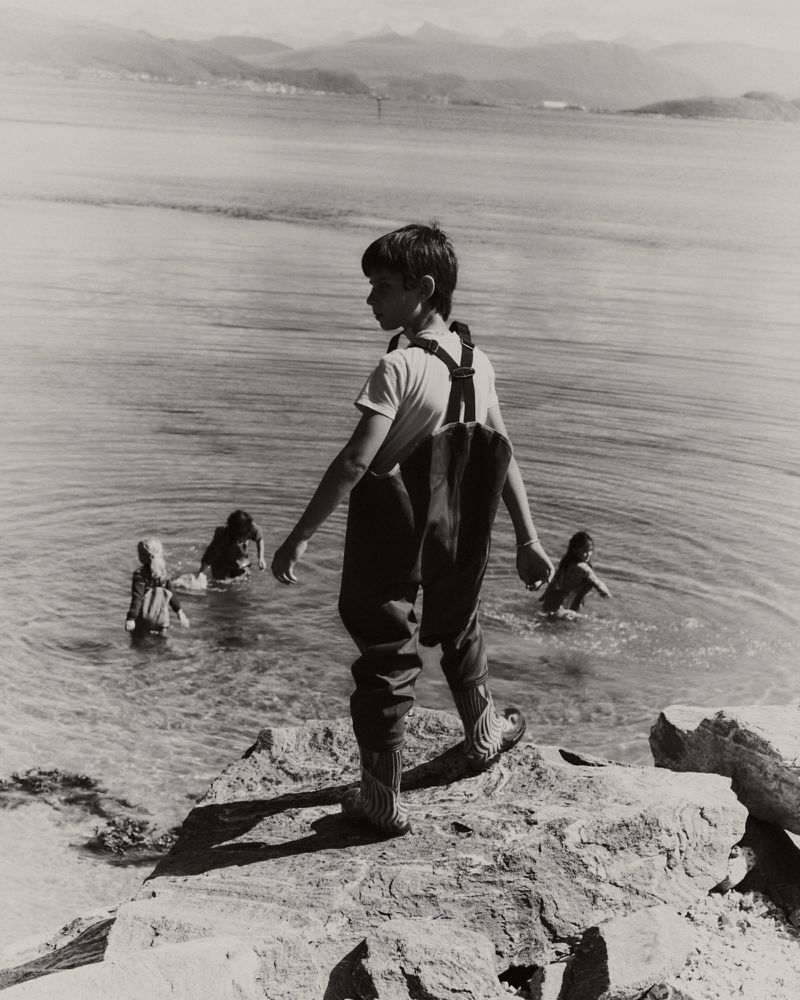 Young boy standing on rocky shore looking out toward water with other children wading in the background under an overcast sky.