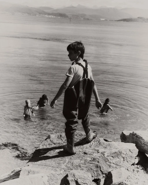 Young boy standing on rocky shore looking out toward water with other children wading in the background under an overcast sky.
