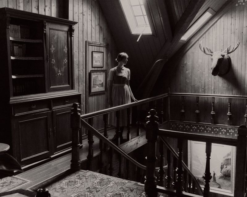 Woman standing on a wooden mezzanine overlooking a staircase inside a rustic cabin with wood-paneled walls and a mounted moose head.