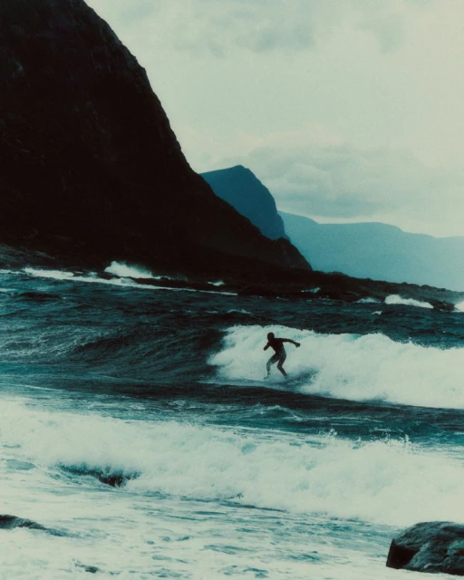 Surfer riding a wave in rough ocean waters beneath steep dark coastal cliffs in Norway