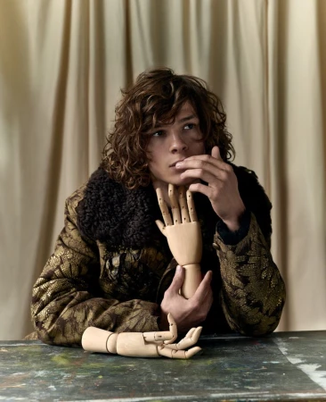 Young man with curly brown hair wearing a patterned jacket posing with wooden artist hands for ICON Magazine France