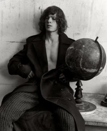 Portrait of young man with curly hair wearing an open dark coat and ribbed trousers beside a globe on a table
