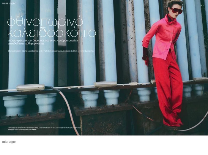 Model wearing a pink jacket and red trousers standing against large industrial pipes for Vogue Greece